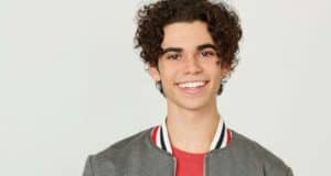 Curly-haired guest in gray jacket and red shirt smiles warmly, evoking beloved Disney Channel star Cameron Boyce.
