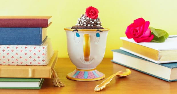 A Chip mug cupcake display, surrounded by books, a rose, and golden spoon, evokes Beauty and the Beast magic on a wooden table.