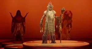 A Lion King performer in stunning tribal costume wows the stage at Disneyland Paris, staff in hand against a vivid orange backdrop.