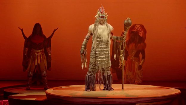 A Lion King performer in stunning tribal costume wows the stage at Disneyland Paris, staff in hand against a vivid orange backdrop.