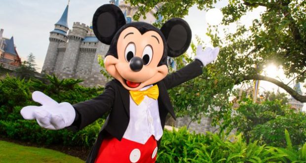 Mickey Mouse welcomes guests with open arms in front of the iconic Disney World castle, surrounded by lush greenery.