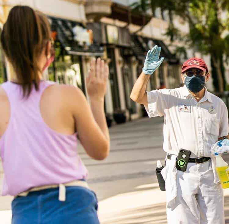 A masked Disney Cast Member gives a high five to a guest outside on a sunny day at the park.