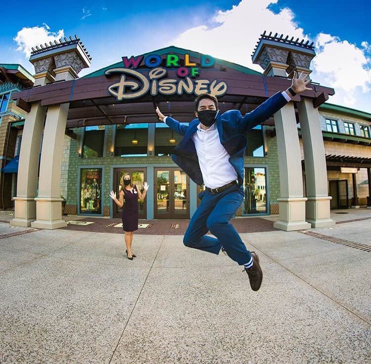 A guest in a suit jumps for a photo op in front of the World of Disney at Disney Springs, with another guest enjoying the moment.