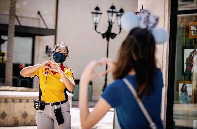 A Cast Member and a Disney Springs visitor form heart shapes with their hands, both sporting face masks at the outdoor venue.