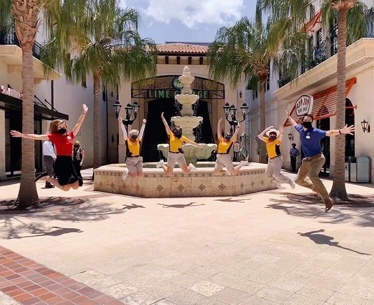 Six Disney fans in masks leap mid-air by the iconic fountain at Disney World, palm trees swaying under a bright blue sky.