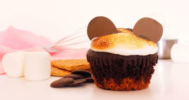 A chocolate cupcake with toasted marshmallow frosting and two round chocolate pieces on top, resembling bear ears, sits on a white surface with marshmallows, graham crackers, and chocolate pieces in the background.