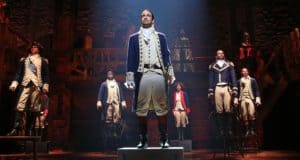 The cast of "Hamilton" performs on Broadway in vibrant colonial attire, with dramatic lighting—now streaming on Disney+.