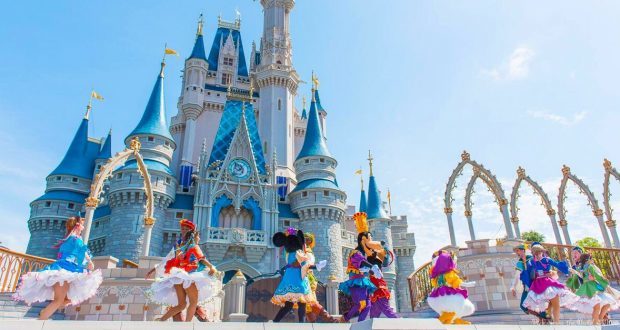 Disney performers in vibrant costumes dance before Cinderella Castle under sunny skies at Walt Disney World.