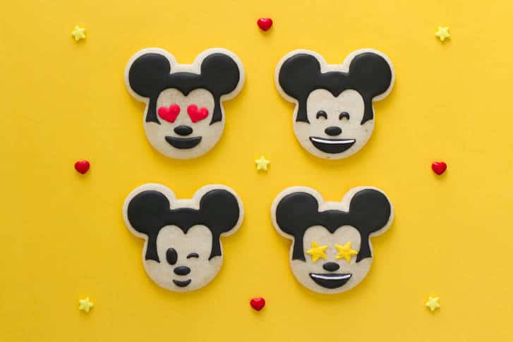 Four Mickey Mouse emoji cookies on a yellow background with hearts and stars—Disney magic for fans and party inspo!.