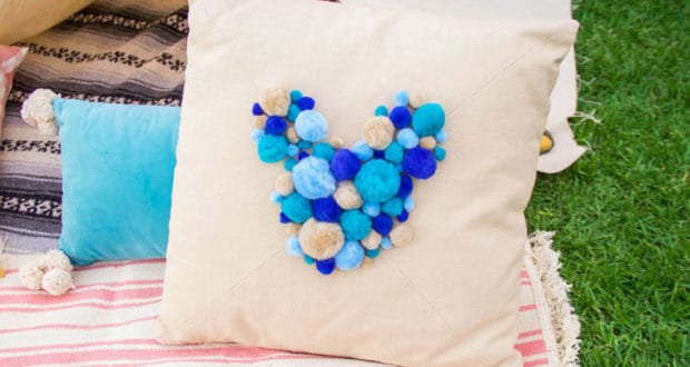 Bear-shaped DIY pillow with blue and tan pom-poms, styled like theme park merch, sits on playful blankets outside on the grass.