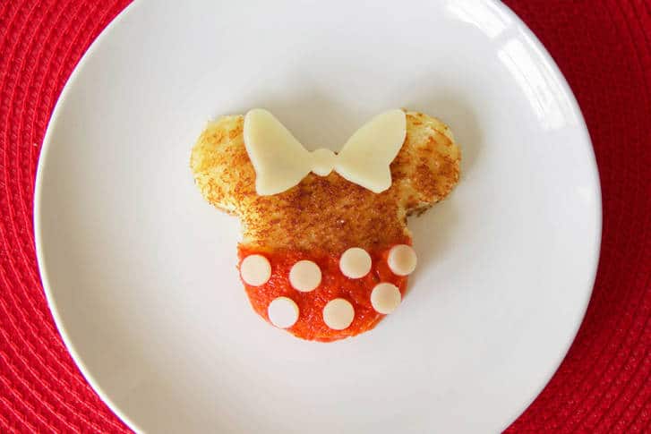 Grilled cheese toast styled like Minnie Mouse with red jam, cheese dots, and bow, served on a plate—perfect for Disney parks fans.