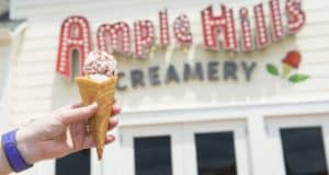 A hand holds an ice cream cone with sprinkles in front of the Ample Hills Creamery sign. The sign features retro-style lettering with lights and a small ice cream cone graphic. The background is the cream-colored exterior of the building.