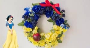 Snow White wreath with blue and yellow blooms, red bow, and apple beside Snow White artwork—magical Disney Parks-inspired decor!.