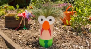 A whimsical bottle planter with a cartoon face and grassy hair brightens the garden, surrounded by vibrant blooms and props.
