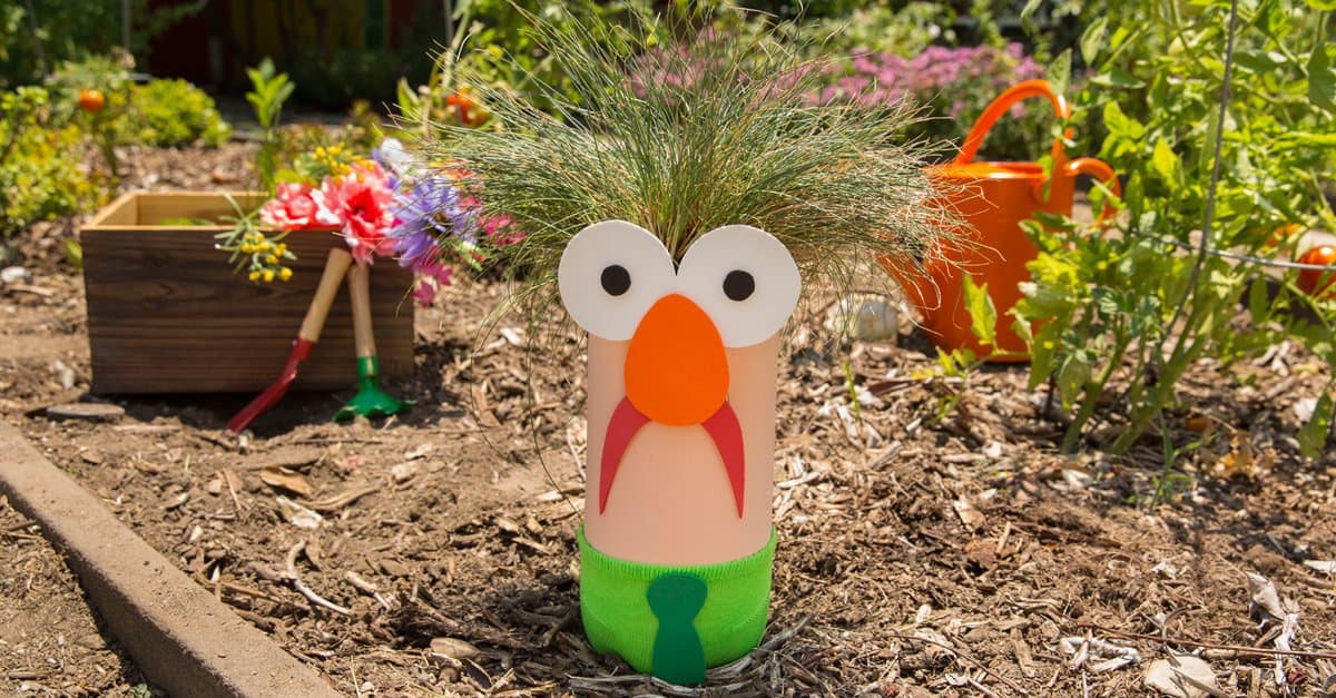 A whimsical bottle planter with a cartoon face and grassy hair brightens the garden, surrounded by vibrant blooms and props.