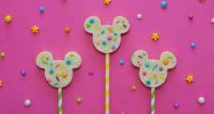 Mickey-shaped lollipops with colorful sprinkles pop against a pink, starry Disney-inspired background, perfect for park festivities.