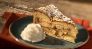 A magical apple cake slice dusted with powdered sugar, topped with whipped cream, served on a dark plate for theme park guests.
