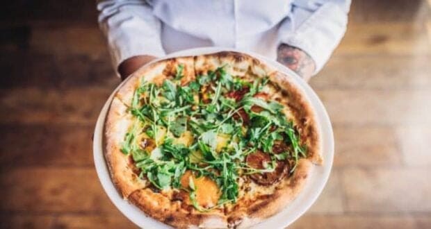 A person holding a freshly baked pizza topped with arugula and assorted vegetables on a thin, crispy crust, standing on a wooden floor. The pizza is presented on a white plate.