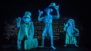 The Hitchhiking Ghosts from Haunted Mansion pose in front of a dark brick wall, one holding a suitcase, glowing blue.