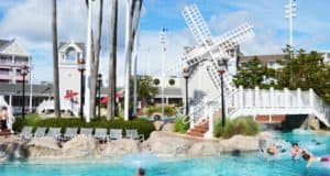 Guests splash in a lively resort pool near a white windmill, rock formations, and themed gray buildings at the entertainment complex.