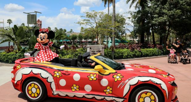 Minnie Mouse waves from a red, polka-dot convertible adorned with flowers, capturing the enchantment of Disney World.