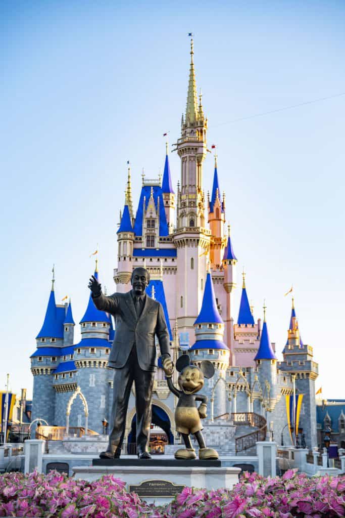 Walt Disney and Mickey Mouse statue stands before Cinderella Castle at Disney World, framed by vibrant pink flowers in bloom.