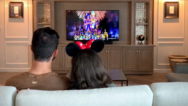Disney fans relax on the couch, enjoying a dazzling fireworks show on TV; one sports iconic Minnie Mouse ears for extra magic.