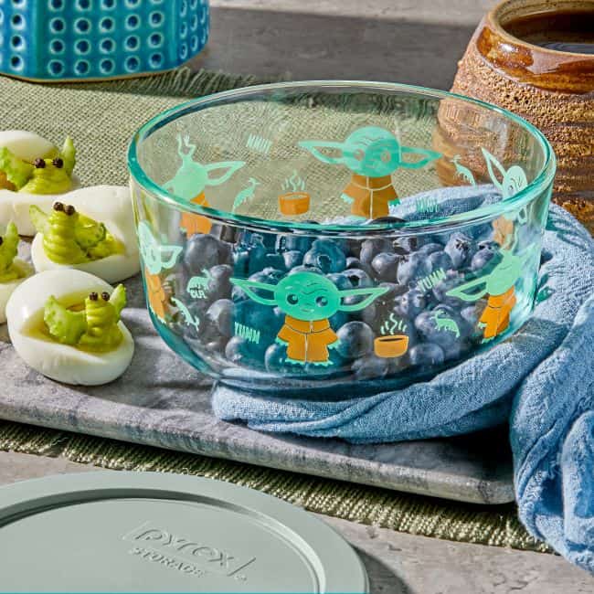 A Star Wars Corelle Collection bowl featuring Baby Yoda is filled with blueberries, set alongside a tray of deviled eggs crafted to look like Baby Yoda—perfect treats for fans visiting Galaxy’s Edge or celebrating Star Wars Day at a themed event.