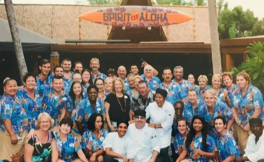 Entertainers in coordinated blue Hawaiian shirts gather beneath the “Spirit of Aloha” sign at Disney's Polynesian Village Resort, ready to welcome guests with island-inspired performances.