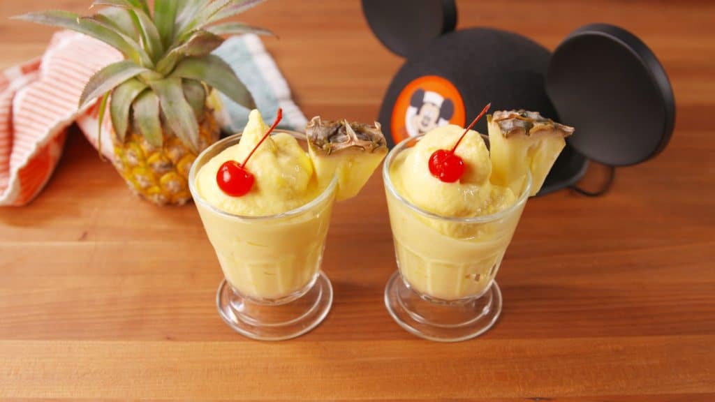 Two glasses of Dole Whip topped with cherries and fresh pineapple slices sit in front of a classic Mickey Mouse ears hat, capturing the iconic Disney Parks treat. This copycat pineapple whip recipe channels the sweet, refreshing flavors guests love at Disney World, bringing a bit of that signature magic straight to your celebration.