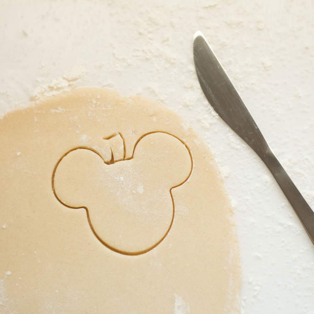Mickey Mouse-shaped cookie dough ready to bake, with a butter knife on a floured surface—just the thing for Disney fans looking to bring some theme park magic home, especially when paired with your favorite Pumpkin Spice Mickey Hot Cocoa.