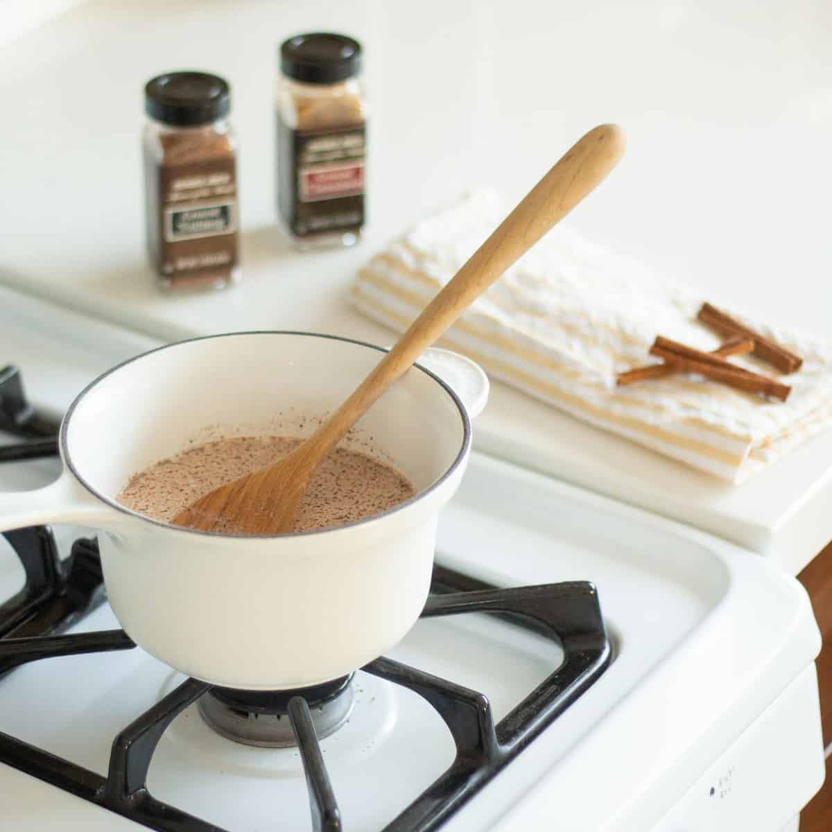 A festive white pot with a wooden spoon stirs rich cocoa on the stovetop, showcasing the Pumpkin Spice Mickey Hot Cocoa Recipe inspired by Disney Parks, surrounded by autumn spices and soft towels for that magical at-home Disney treat experience.