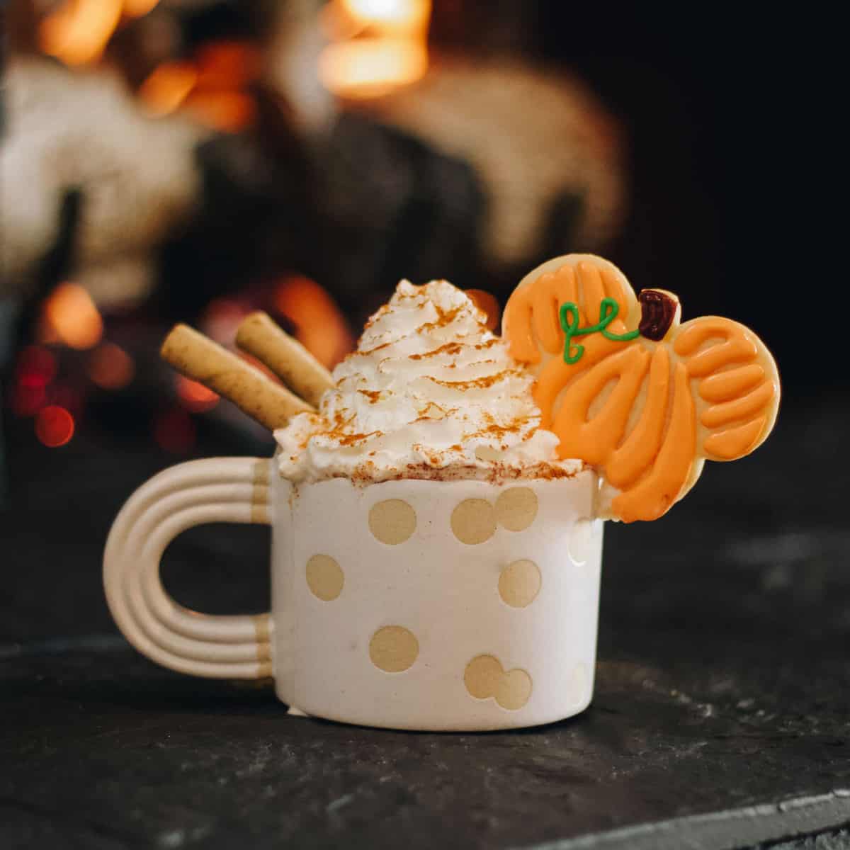 A whimsical mug brimming with whipped cream, crisp wafer sticks, and a pumpkin-shaped Mickey cookie captures the magic of Disney’s Pumpkin Spice Hot Cocoa, set aglow by the warm hearth—bringing a dash of theme park coziness right to your cup.