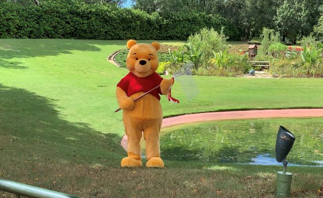 Winnie the Pooh stands by a scenic pond in a lush green park, charming guests with a butterfly net—just like meeting this beloved character at Walt Disney World or EPCOT.