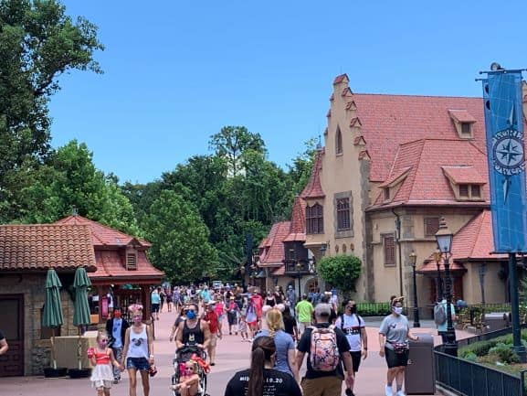 Guests stroll down a scenic walkway at Walt Disney World, surrounded by vibrant red-roofed resort buildings and lush trees beneath a bright blue sky.