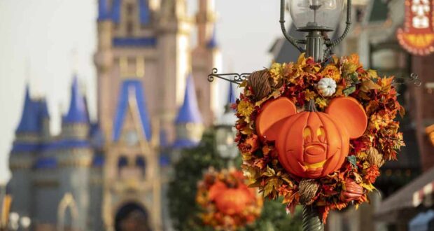 A Mickey Mouse-shaped pumpkin wreath decorated with autumn leaves hangs on a lamppost; Cinderella Castle is blurred in the background, creating a festive Halloween scene at a Disney park.