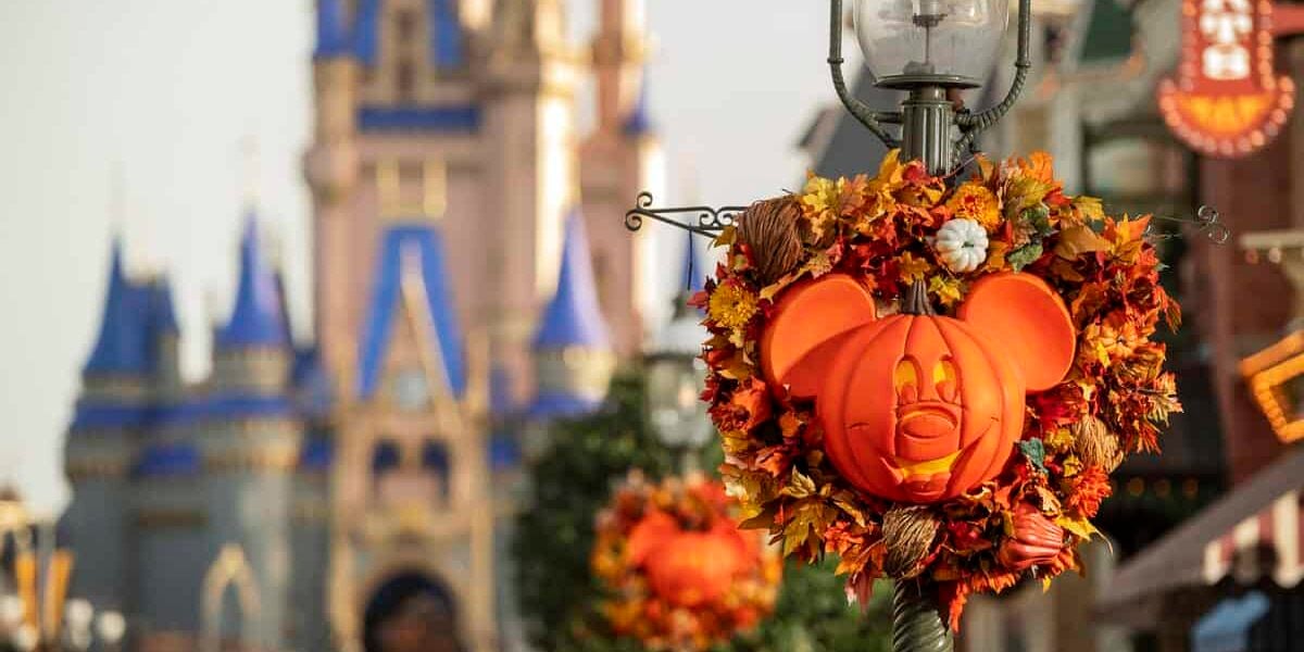 A Mickey Mouse-shaped pumpkin wreath decorated with autumn leaves hangs on a lamppost; Cinderella Castle is blurred in the background, creating a festive Halloween scene at a Disney park.