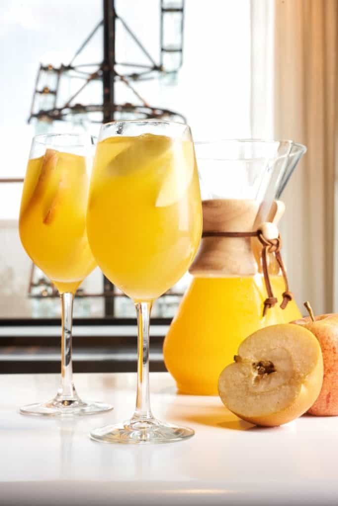 Two glasses of golden cocktail with ice and apple slices are ready to sip beside a carafe and freshly sliced apples, capturing the flavors of Disney Springs’ Fall Specialty Cocktails for guests seeking festive tastes during their visit.