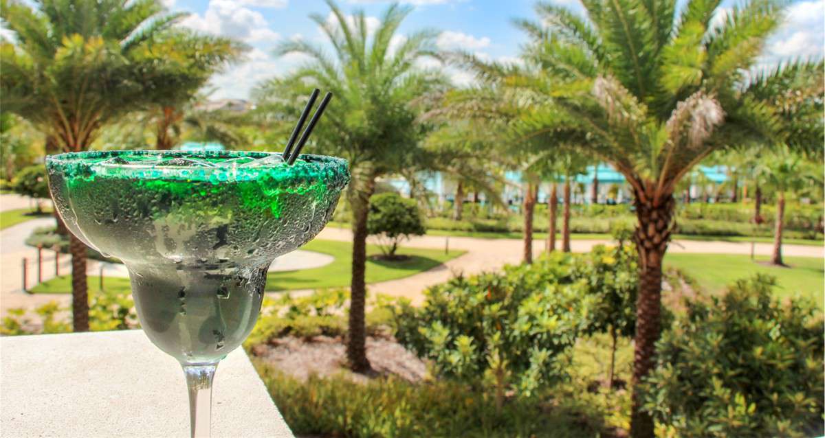 A vibrant green cocktail with straws sits ready to enjoy, set against lush palm trees and a sun-drenched garden at Margaritaville Resort Orlando, radiating ultimate vacation vibes for theme park goers.