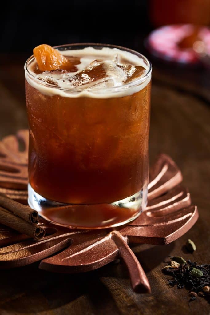 A dark, refreshing iced cocktail with an orange garnish rests on a leaf-shaped coaster alongside cinnamon sticks, ready to delight guests as part of the Fall Specialty Cocktails lineup at Disney Springs.