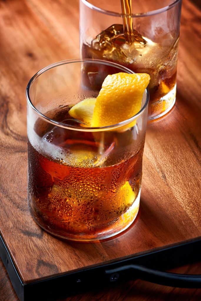 Enjoying two glasses of whiskey on the rocks with orange peel garnish, served on a rustic wooden tray at Disney Springs—one glass being poured to kick off signature Fall Specialty Cocktails in true Disney style.
