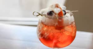 A vibrant orange cocktail with ice, topped with creamy foam and crowned by a fruit garnish, sends wisps of theatrical smoke into the air as it sits on a rustic wooden table—a showstopping drink that looks right at home as the whimsical signature cocktail at Margaritaville Resort Orlando’s Professors Poison Apple.
