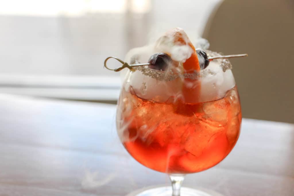 A vibrant red cocktail bubbling with smoke and topped with foam and garnish sits in a round glass on a table, evoking the magical flair of the Professors Poison Apple drink found at Margaritaville Resort Orlando—perfect for fans seeking themed drinks during their resort adventure.