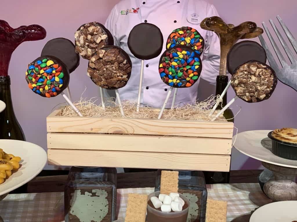 Chocolate-dipped cookie pops topped with vibrant candies and crunchy nuts in a rustic wooden box, an irresistible EPCOT treat after a day of adventures; Disney chef nearby, ready to dish out insider tips for your Walt Disney World visit.