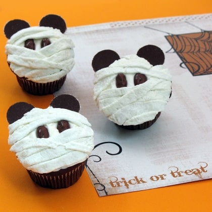 Delight in these festive Mickey Mummy Cupcakes, perfectly themed for Halloween at Disney Parks. Each cupcake is decorated to look like a mummy with chocolate Mickey ears, displayed on an orange backdrop and festive "trick or treat" paper—capturing that playful Disney magic for the season.