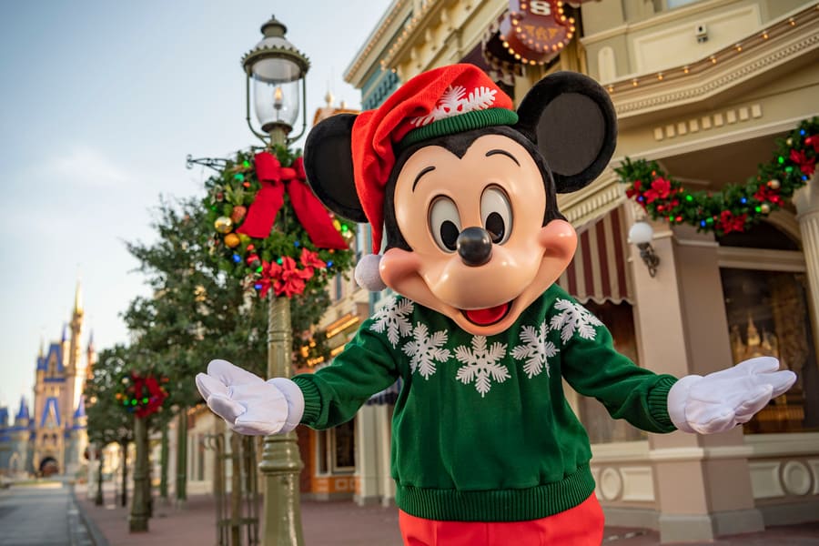 Mickey Mouse sports a green holiday sweater and red hat as he greets guests on a festively decorated street adorned with Christmas garlands, adding extra Disney magic to the seasonal atmosphere—just like the enchanting Character Cavalcades at Walt Disney World.