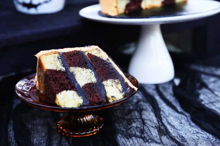 A slice of black and white checkerboard cake with chocolate drizzle, served on a glass plate that channels Tim Burton’s Nightmare Before Christmas magic, with a cake stand in the background setting the stage for a delightfully spooky dessert at a themed entertainment venue.