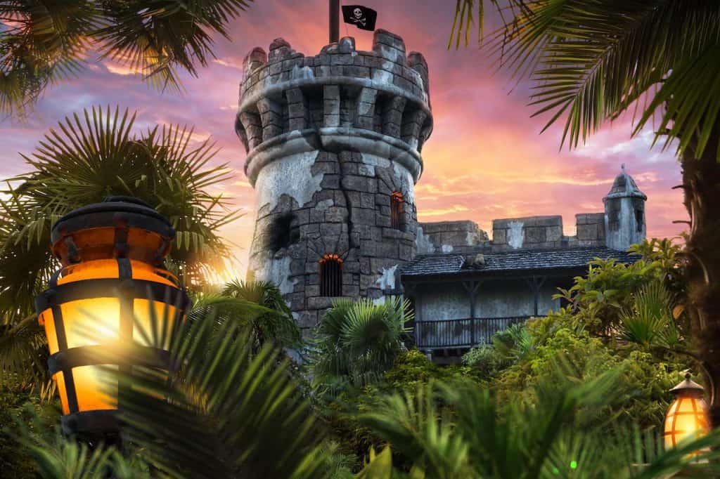 Pirate lookout tower flying a black flag, surrounded by swaying palm trees and glowing lanterns, illuminated by a dramatic sunset and vibrant skies—the perfect backdrop for an adventure at a pirate-themed attraction or live-action show reminiscent of Pirates of the Caribbean.