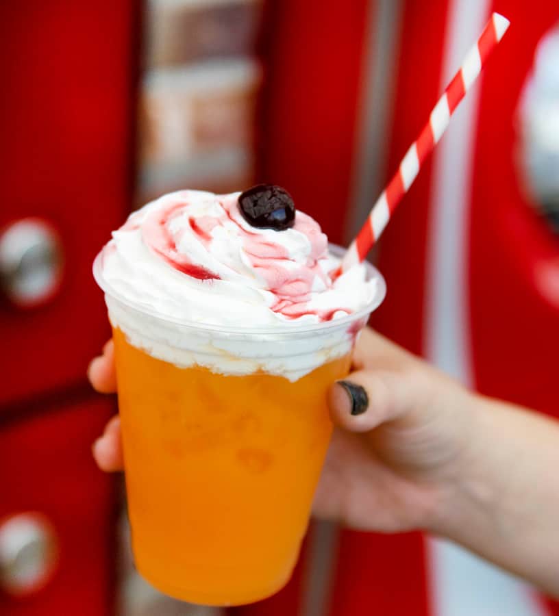 An orange specialty drink topped with whipped cream and a cherry, served with a striped straw in a clear plastic cup at Disney Springs in Walt Disney World Resort.