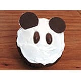 A whimsical cupcake styled like a panda face with white icing, cookie ears, and chocolate candy eyes rests on a wooden counter—an irresistible treat for visitors at the theme park’s Halloween celebration, combining adorable design with sweet theme park magic.
